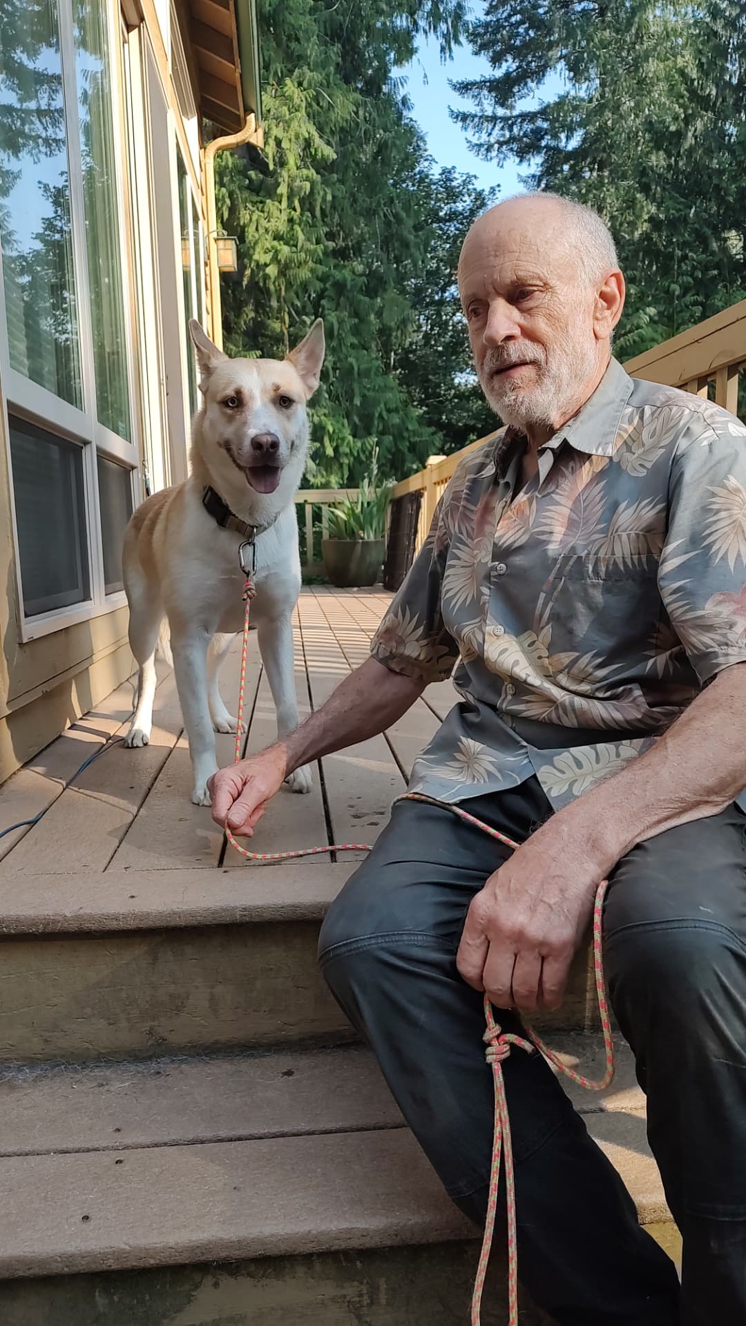 An older man sitting on wooden porch steps holding a leash beside a happy cream and tan rescue dog